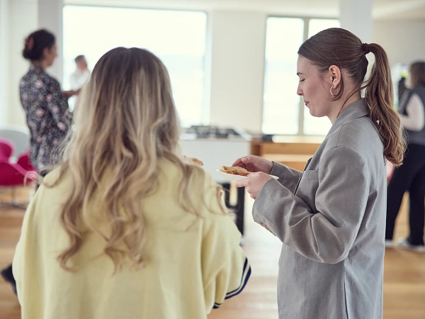 Zwei Frauen stehen in einer modernen, hellen Büroumgebung und unterhalten sich, während sie Snacks auf Tellern halten. Im Hintergrund sind weitere Personen zu sehen, die sich in der lockeren Atmosphäre bewegen.