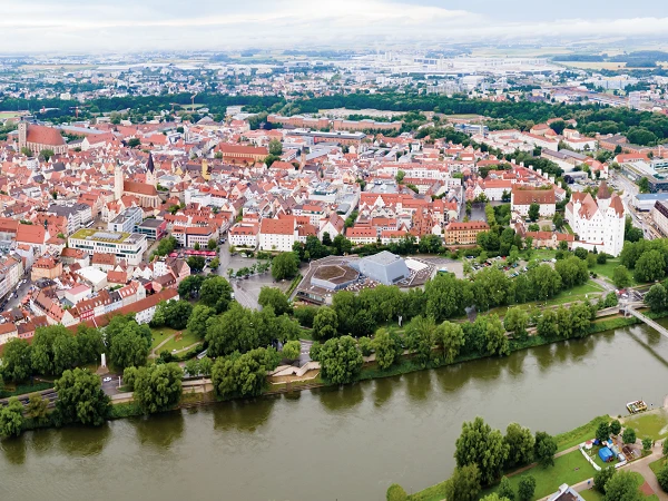 Luftaufnahme einer Stadt mit roten Dächern, umgeben von grünen Bäumen und einem Fluss, der sich durch die Landschaft schlängelt. Im Hintergrund sind moderne Gebäude und eine weite Landschaft zu sehen.