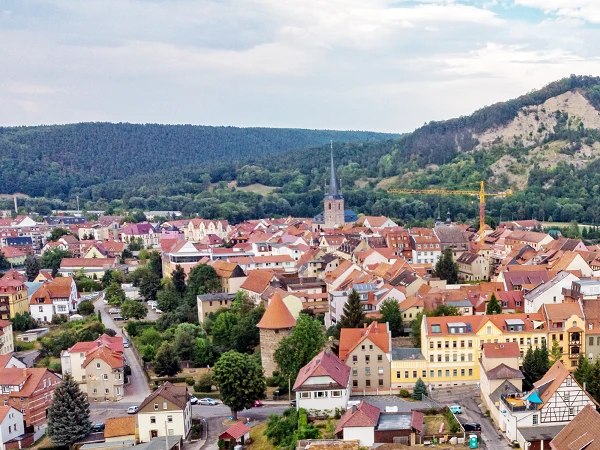Panoramaansicht einer kleinen Stadt mit roten Dachziegeln, umgeben von bewaldeten Hügeln und einer Kirche im Zentrum. Der Himmel ist teilweise bewölkt und im Hintergrund ist eine Baustelle mit einem Kran zu sehen.
