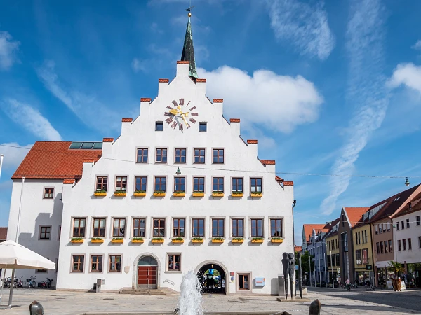 Das Rathaus mit einem markanten, weiß getünchten Giebel und einem Ziffernblatt an der Fassade steht auf einem Platz, umgeben von weiteren Gebäuden. Ein Brunnen mit Wasserstrahl befindet sich im Vordergrund, und der Himmel ist klar mit einigen Wolken.