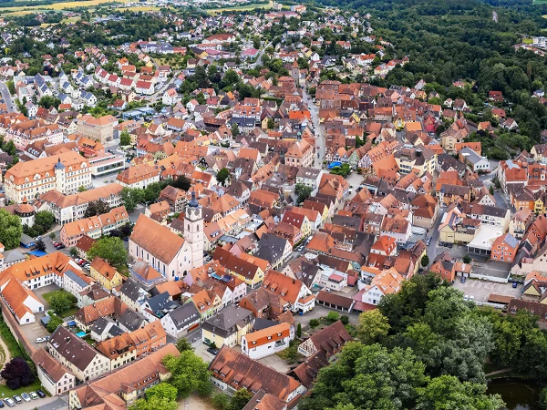 Eine Luftaufnahme einer malerischen Stadt mit zahlreichen roten Dächern und einer historischen Kirche im Zentrum, umgeben von grünen Wäldern und Feldern.