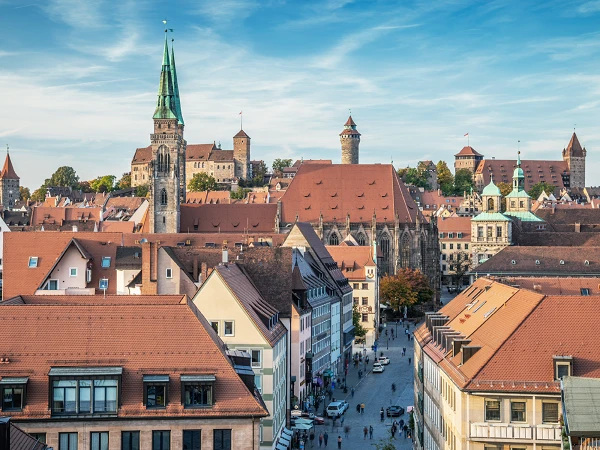Blick auf die Altstadt von Nürnberg, mit roten Ziegeldächern und historischen Gebäuden, darunter der Hauptmarkt und die Kirche St. Sebald, umgeben von Hügeln und einem klaren blauen Himmel.