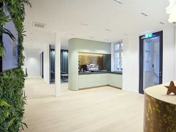 Ein moderner Innenraum mit hellen Holzböden, der an einer Seite eine grüne Pflanzenwand und an der anderen eine elegante Küchenzeile mit Kaffeemaschine zeigt. Helle Wände und Fenster sorgen für eine freundliche Atmosphäre.