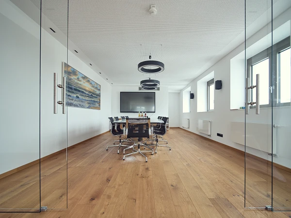 Ein moderner Konferenzraum mit Holzboden, einem großen Tisch in der Mitte, umgeben von Drehstühlen, und einem Bildschirm an der Wand. Große Fenster lassen viel Tageslicht herein, und an einer Wand hängt ein Bild mit einer Landschaft.