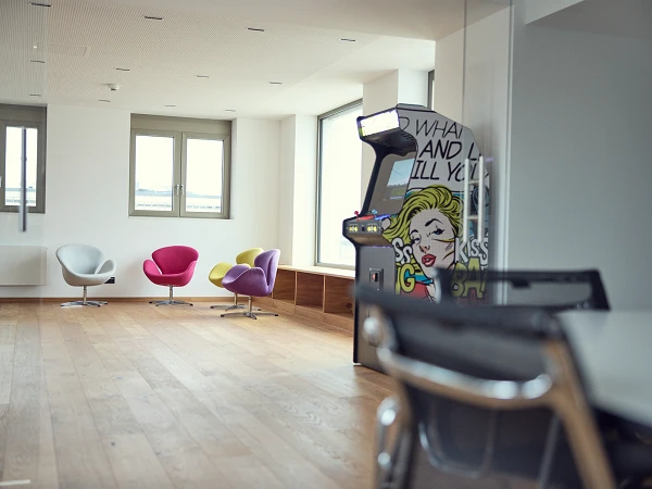 Ein moderner Büroraum mit hellen Wänden und großen Fenstern, in dem ein stilvoller Tisch mit Stühlen, ein paar farbige Sitzmöbel in Pink und Weiß sowie ein Retro-Spiel-Automat mit einem bunten Comic-Design stehen.