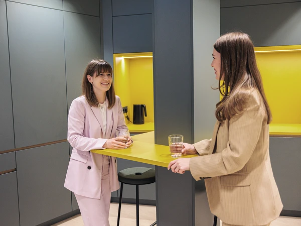 Zwei Frauen stehen in einer modernen Küchenecke mit gelben Akzenten, während sie miteinander sprechen und jeweils ein Glas in der Hand halten.