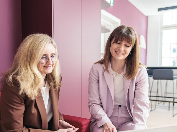 Zwei Frauen sitzen auf einem roten Sofa in einem modernen Büro und betrachten gemeinsam einen Laptop. Eine Frau trägt eine braune Blazerjacke und hat lockiges Haar, während die andere eine hellrosafarbene Anzugjacke trägt und glattes Haar hat. Beide lächeln und wirken konzentriert. Im Hintergrund sind sichtbare Büromöbel und eine Fensterfront.