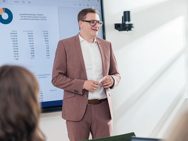 Ein Mann in einem hellen Anzug steht in einem Besprechungsraum und präsentiert vor einer Gruppe von Personen, während im Hintergrund ein Bildschirm mit Diagrammen und Daten sichtbar ist.