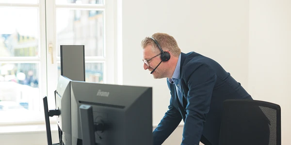 Ein Mann steht an einem Schreibtisch mit mehreren Monitoren und trägt ein Headset. Er scheint in ein Gespräch vertieft zu sein und schaut auf die Bildschirme.