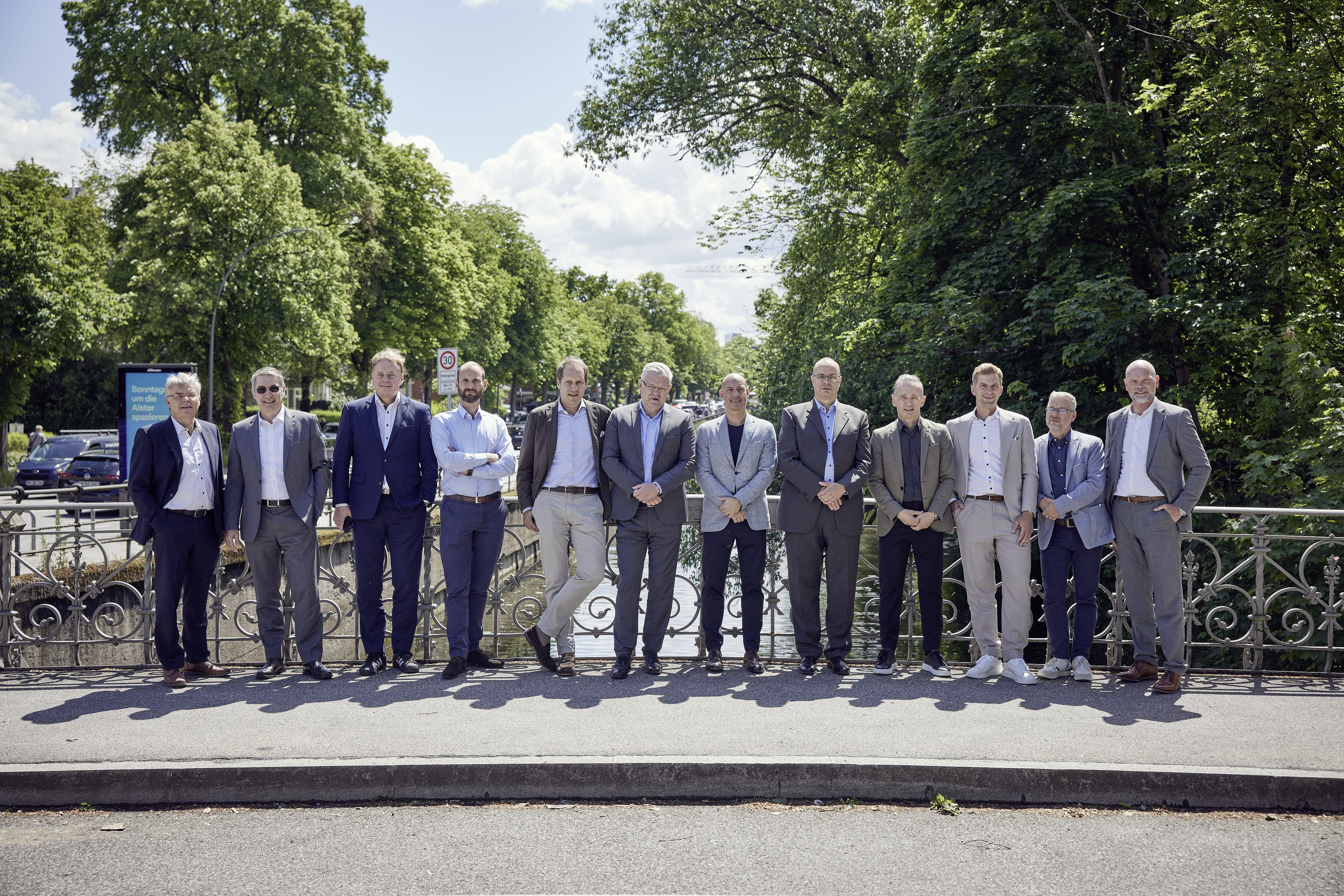 Eine Gruppe von zwölf Männern in Anzügen steht auf einer Brücke, umgeben von Bäumen und einem klaren Himmel im Hintergrund. Sie sind in einem entspannten, freundlichen Arrangement aufgestellt.