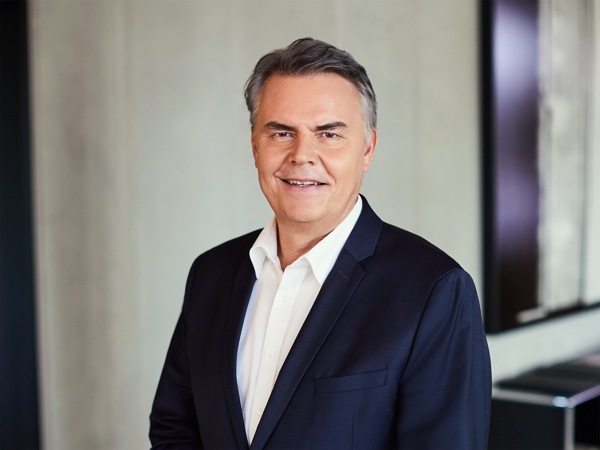 Ein Mann in einem dunklen Anzug und einem weißen Hemd lächelt freundlich in die Kamera, vor einem modernen, minimalistischen Hintergrund.