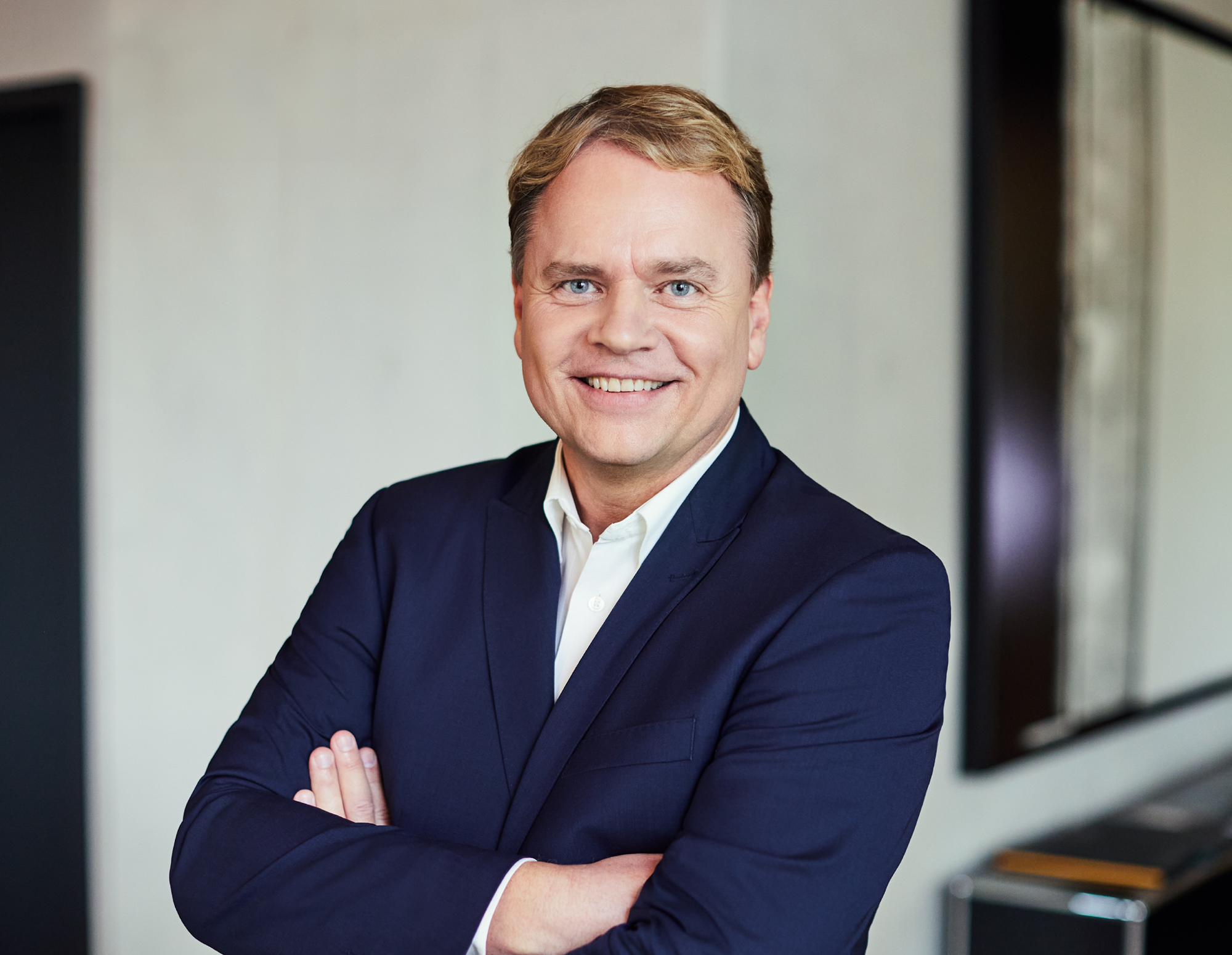 Ein gut gekleideter Mann mit blonden, kurzen Haaren lächelt freundlich in die Kamera, während er mit verschränkten Armen vor einem hellen, modernen Hintergrund steht.
