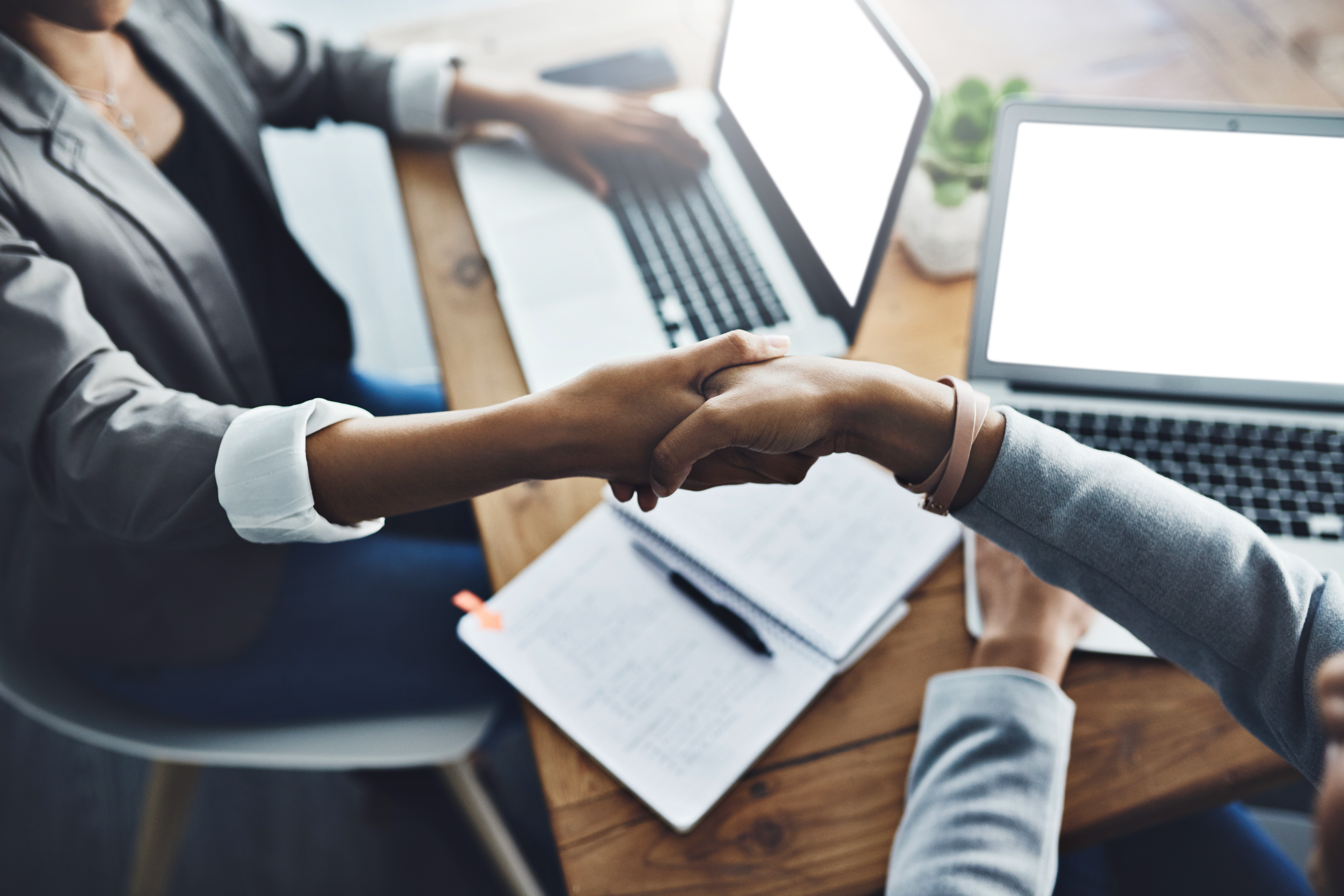 Zwei Personen schütteln sich die Hände über einem Holztisch, auf dem zwei Laptops und Notizblätter liegen.