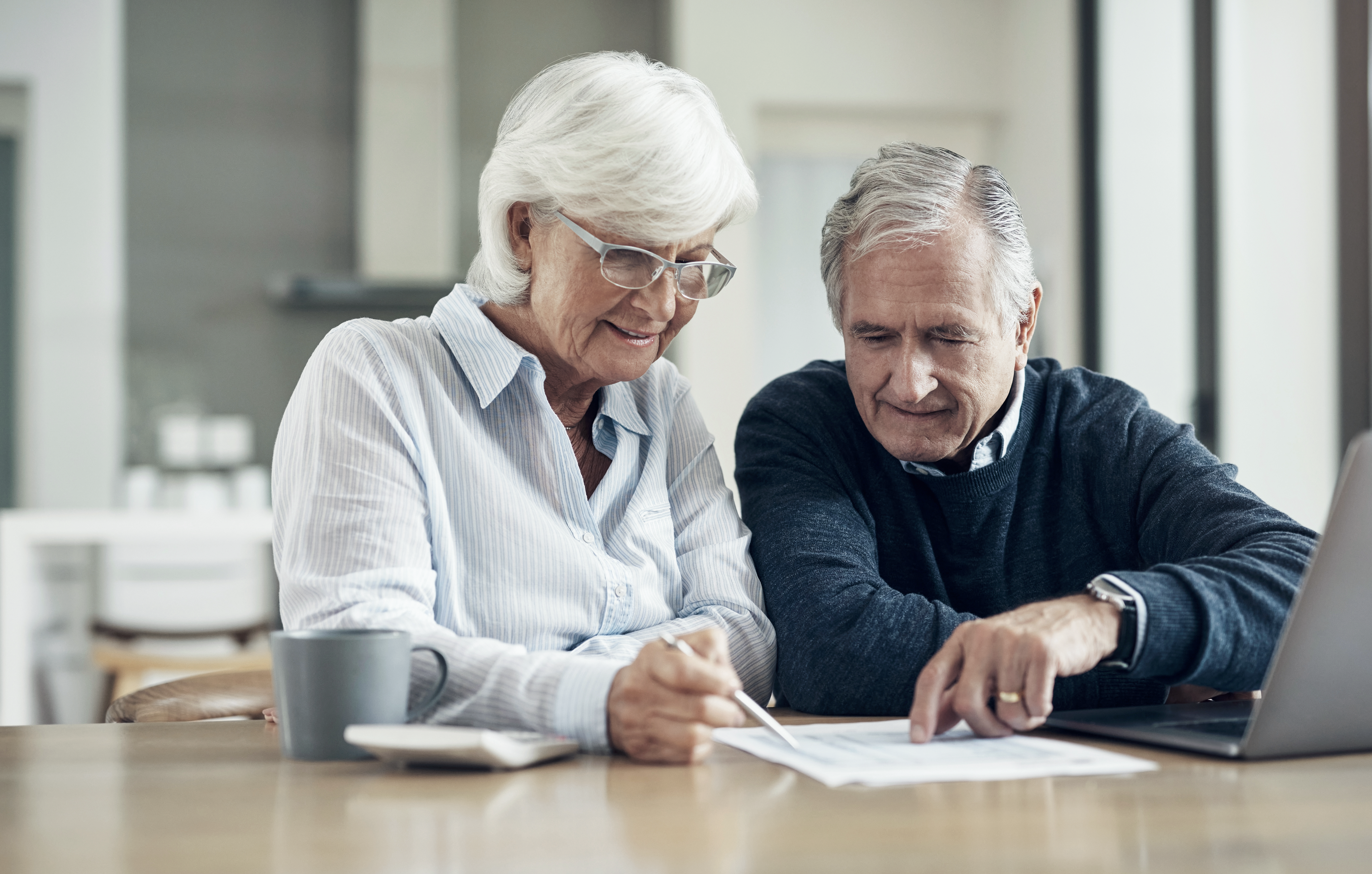 Ein modernes, älteres Paar sitzt am Tisch und geht eine Liste durch, während der Mann am Laptop sitzt.