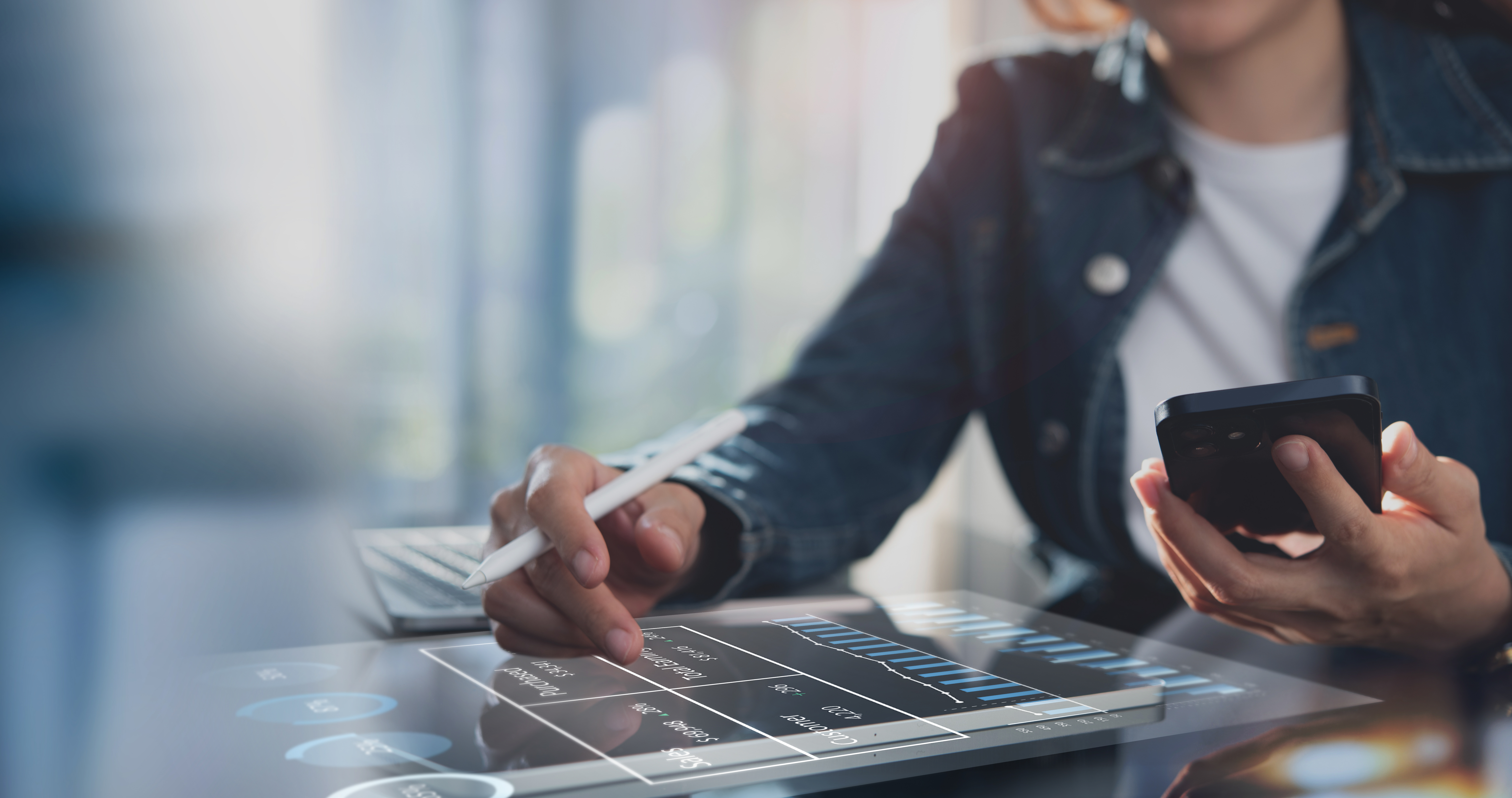 Eine Person in einer Jacke nutzt ein Smartphone in der rechten Hand, während sie mit einem Stylus auf einem Tablet mit grafischen Daten und Diagrammen arbeitet. Im Hintergrund ist ein Laptop sichtbar.