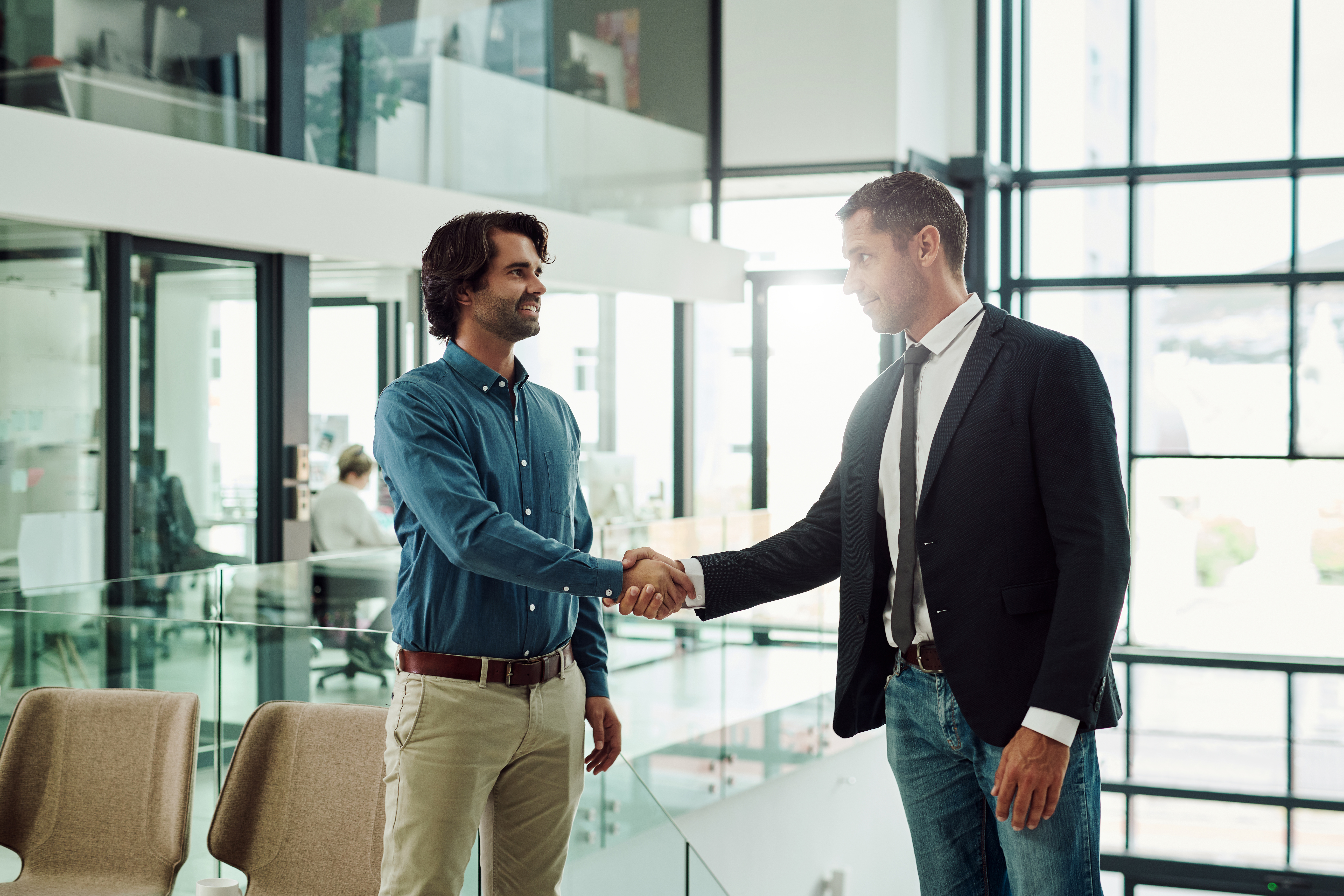Zwei Männer schütteln sich die Hände in einem modernen Büroraum mit großen Fenstern und einem eleganten, offenen Design.