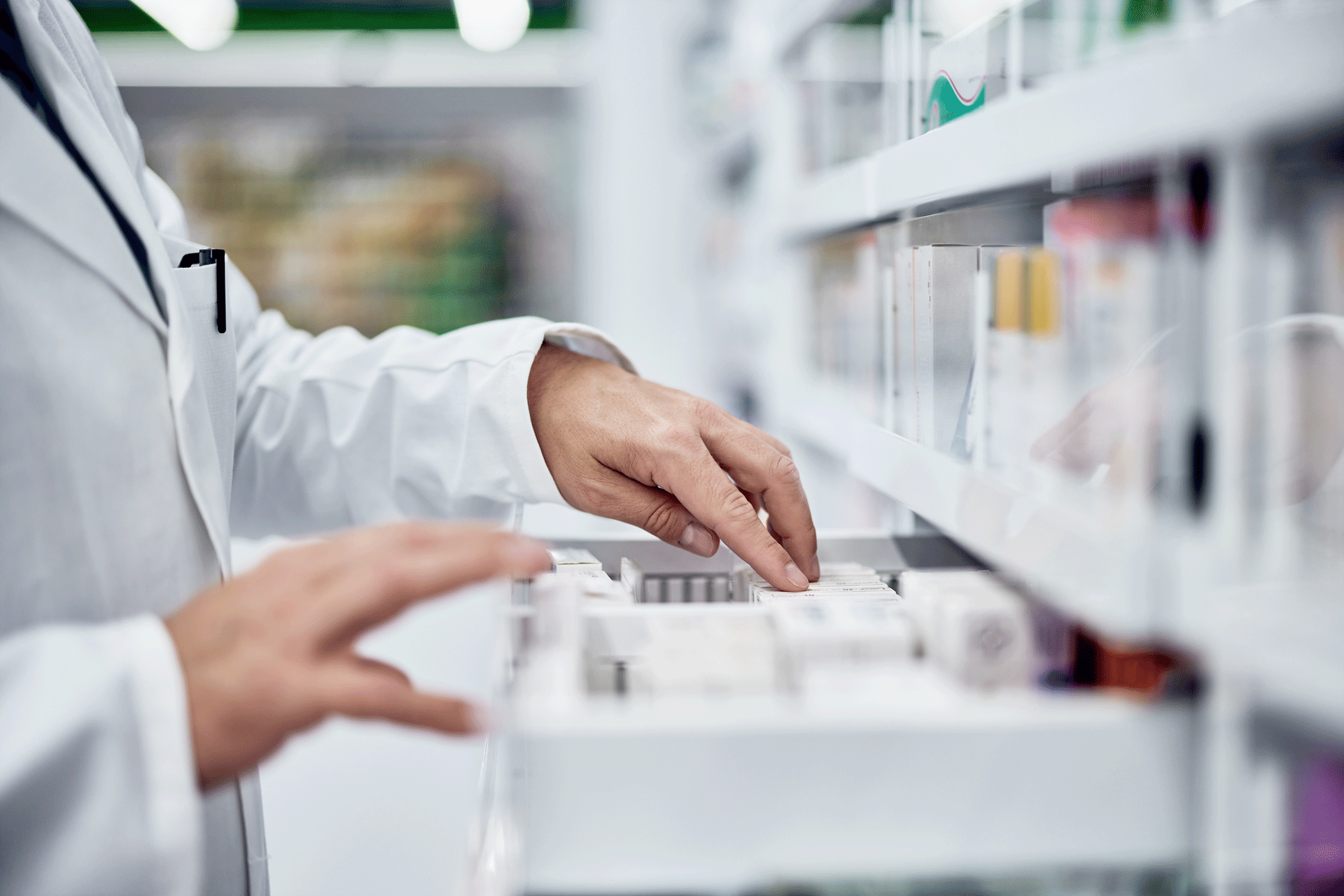 Ein Apotheker in weißem Kittel greift mit der linken Hand nach einem Medikamentenbehälter aus einem Regalsystem, während die rechte Hand auf der Ablage ruhen bleibt, um eine sorgfältige Auswahl zu treffen.