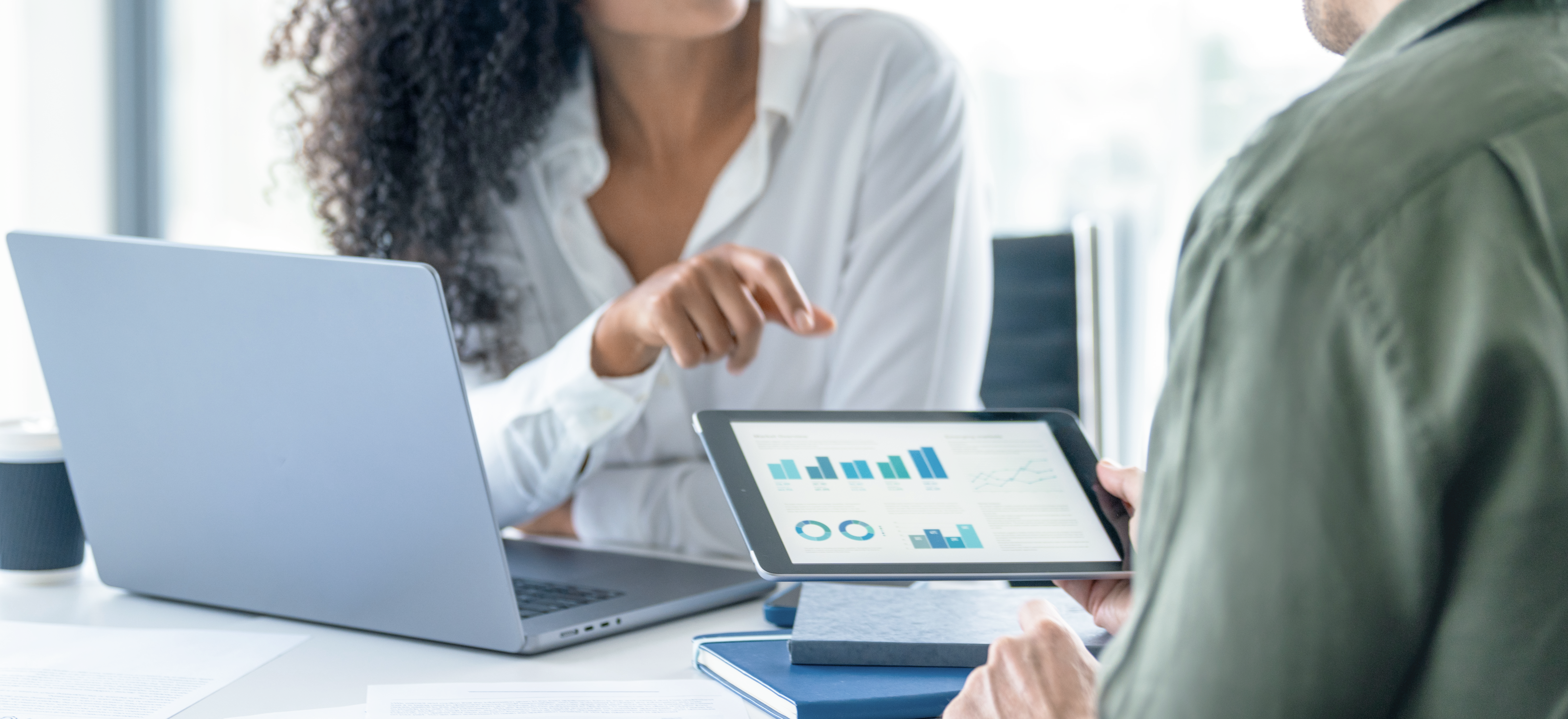 Eine Person mit lockigem Haar zeigt auf eine Grafik auf einem Tablet, während eine andere Person am Tisch sitzt und einen Laptop vor sich hat, in einem modernen Büro.