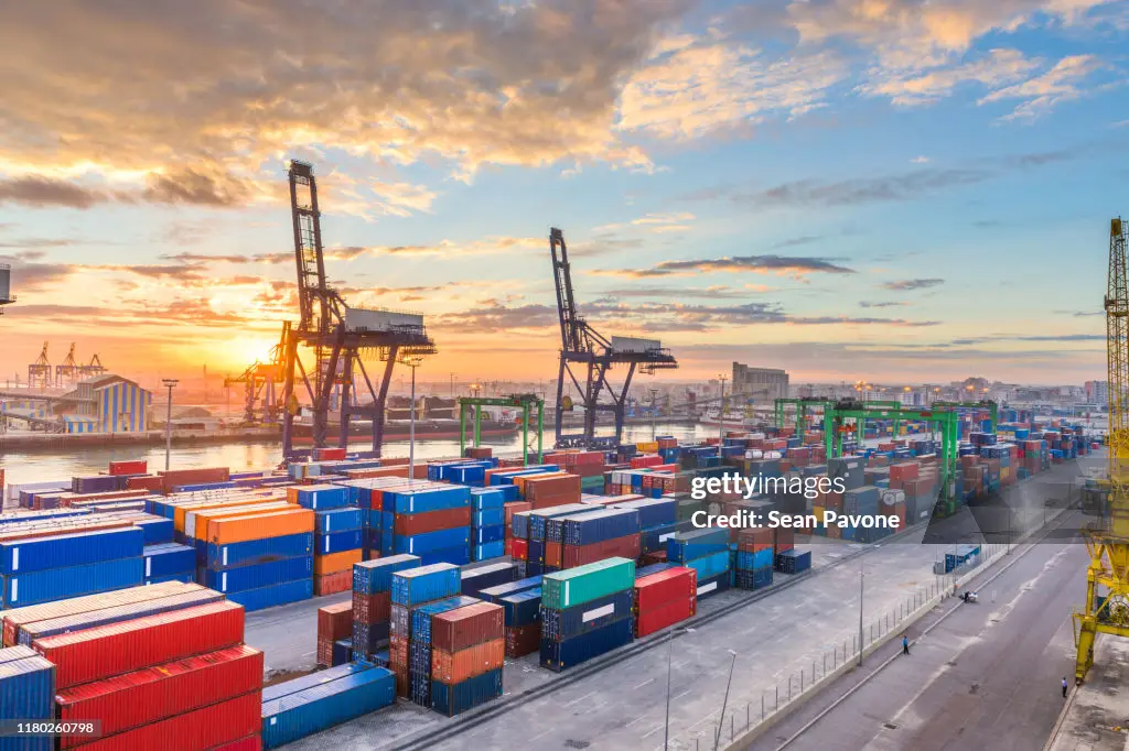 Hafen mit mehreren großen Kränen, die Container bewegen, während die Sonne am Horizont aufgeht und den Himmel in warmen Farben erleuchtet. Containerstapel sind ordentlich auf dem Boden angeordnet.