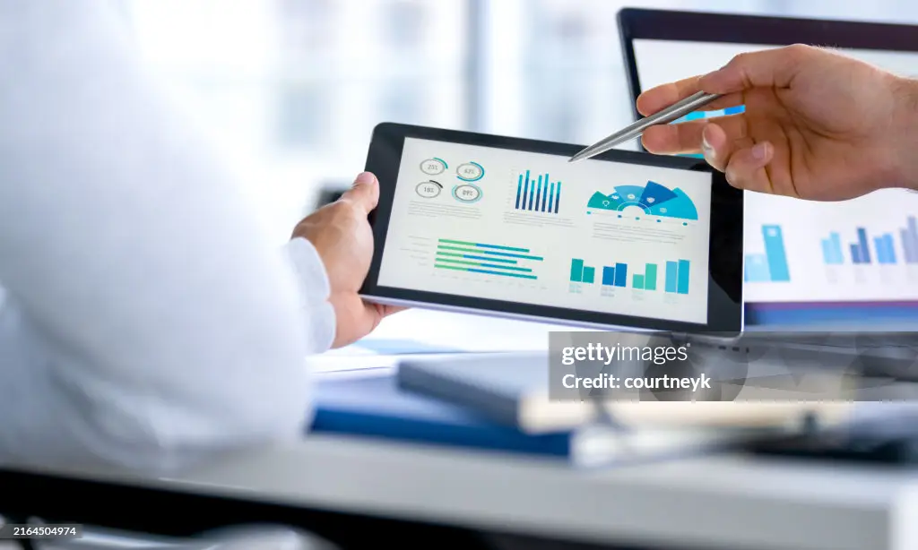 Ein Grafikanalyst zeigt mit einem Stift auf ein Tablet, das verschiedene Diagramme und Datenvisualisierungen anzeigt, während eine andere Person daneben sitzt und zuhört. Im Hintergrund sind weitere Diagramme auf einem Computerbildschirm zu sehen.