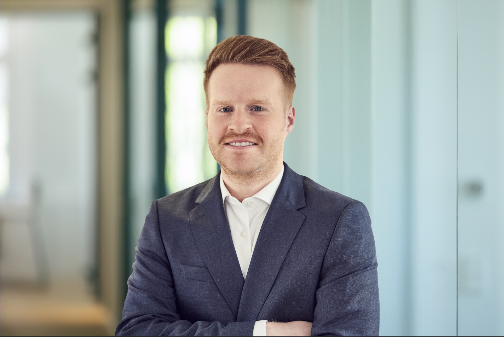Ein Mann mit rotem Haar und Schnurrbart trägt einen dunkelblauen Anzug und steht mit verschränkten Armen vor einer hellen, modern gestalteten Büroeinrichtung.
