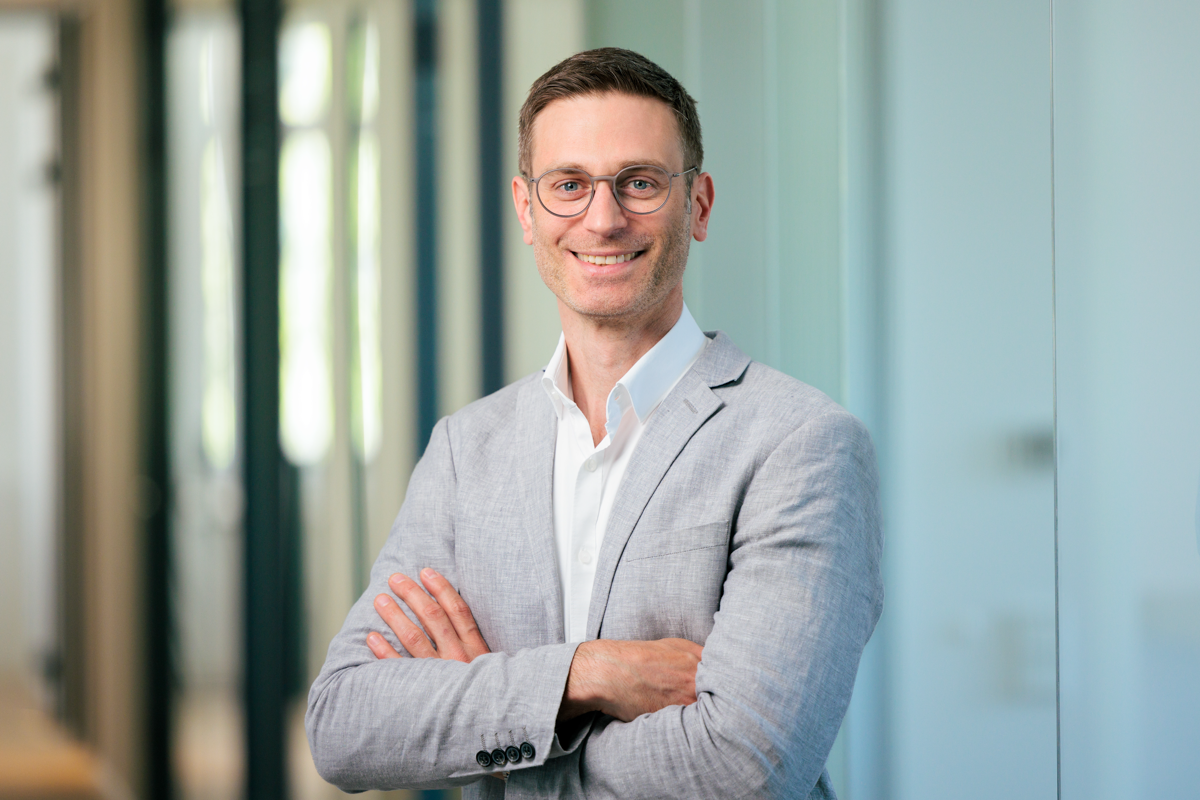 Ein Mann mit Brille trägt einen grauen Blazer und steht lächelnd mit verschränkten Armen vor einem modernen, lichtdurchfluteten Büro.