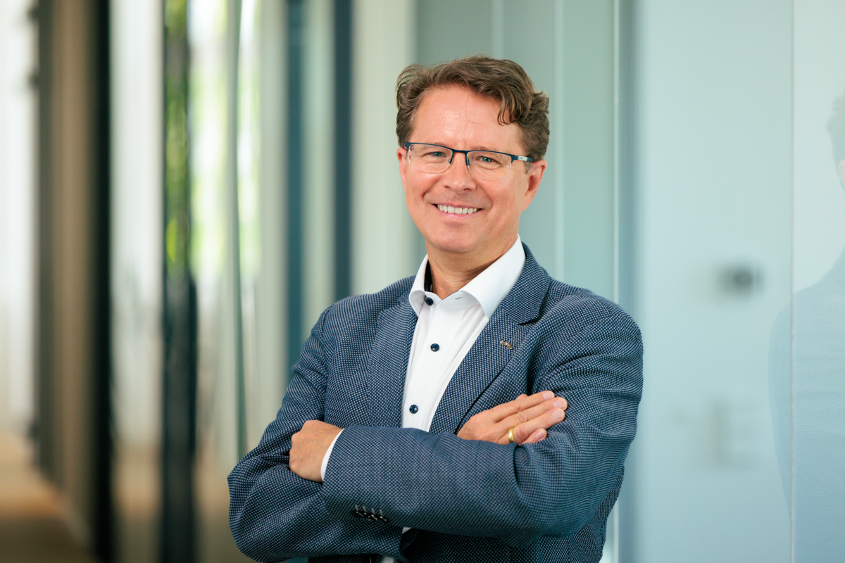 Ein Mann mit Brille und einem lächelnden Gesicht steht mit verschränkten Armen in einem modernen Büroumfeld. Er trägt einen blauen Blazer und ein weißes Hemd mit Knöpfen. Im Hintergrund sind verschwommene Wände und Fenster zu erkennen.