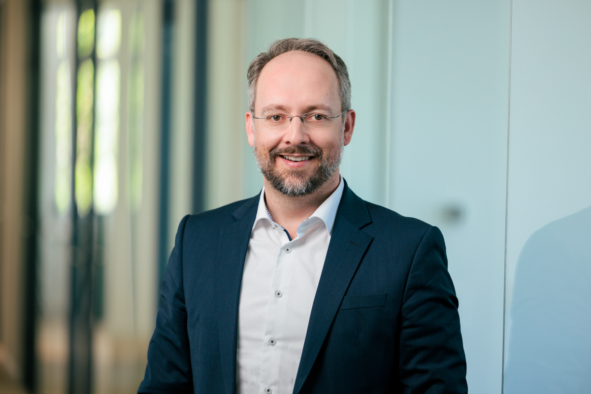 Ein lächelnder Mann in einem dunkelblauen Anzug mit Brille steht in einem modernen Büroraum, umgeben von großen Fenstern und hellen Wänden.