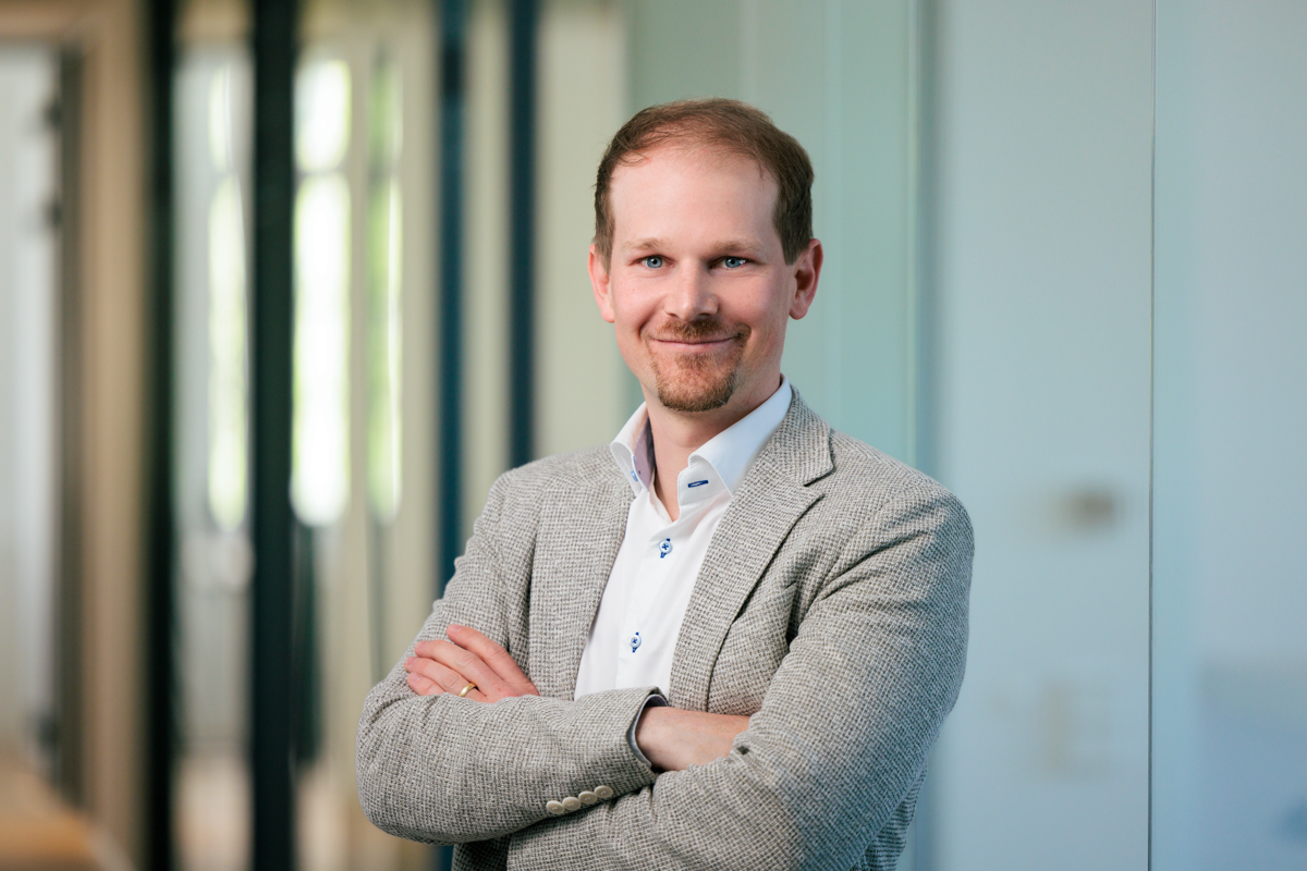Ein Mann mit kurzen Haaren und einem gepflegten Bart steht in einem modernen Büro, lächelt und hat die Arme vor der Brust verschränkt. Er trägt einen hellen Blazer über einem weißen Hemd. Hinter ihm sind unscharfe Büroelemente zu erkennen.