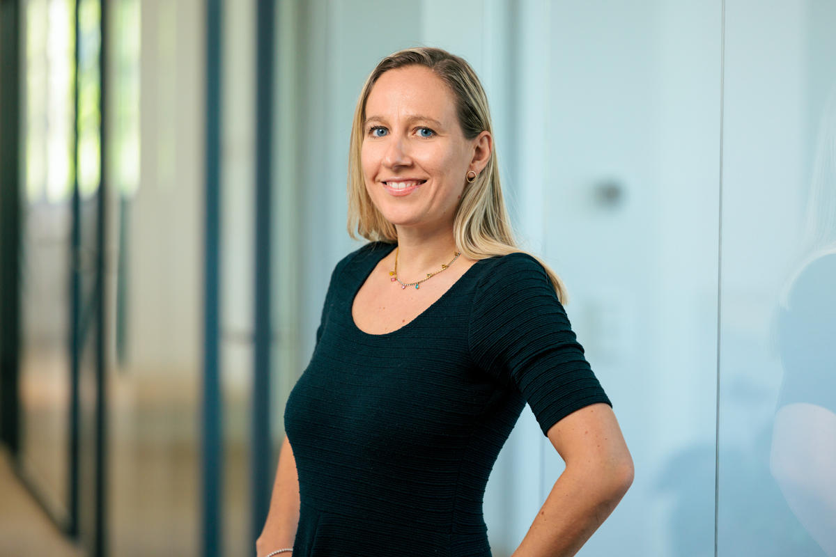 Eine Frau mit langen blonden Haaren trägt ein schwarzes, enges Oberteil und steht lächelnd in einem modernen Büro mit großen Fenstern.