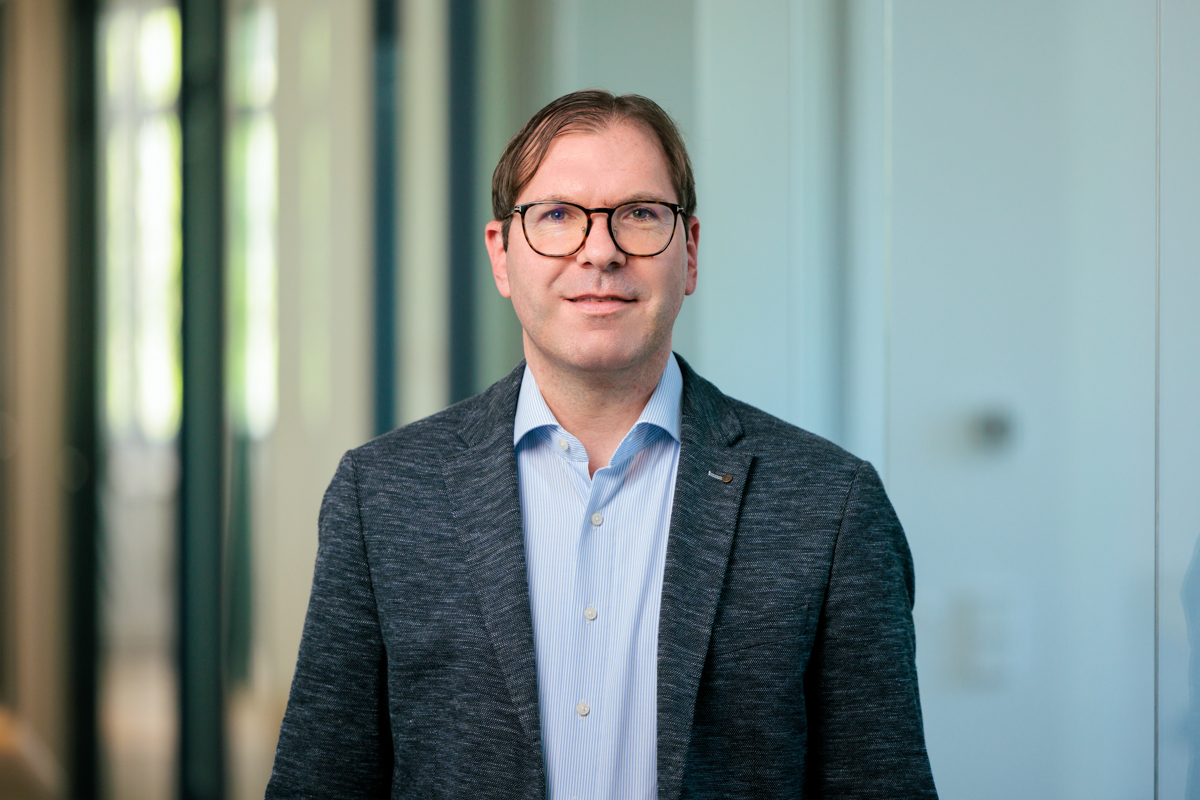 Ein Mann mit Brille, wearing a dunkle Jacke über einem hellen Hemd, steht in einem modernen Büro mit großen Fenstern im Hintergrund.