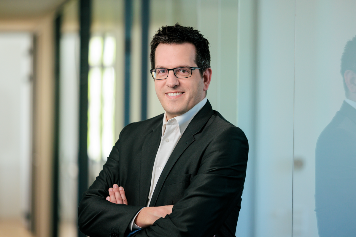 Ein Mann in einem schwarzen Anzug mit einem weißen Hemd und einer dunklen Brille steht in einem modernen Bürogebäude, lächelt und hat die Arme verschränkt. Im Hintergrund sind Glaswände und weiches Licht zu sehen.