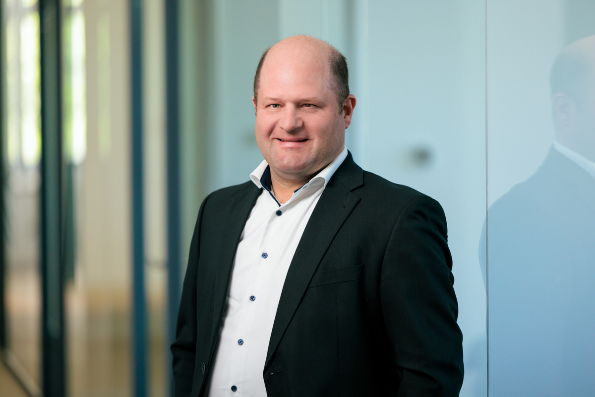 Ein Mann in einem schwarzen Anzug mit einem weißen Hemd steht lächelnd vor einer Glaswand in einem modernen Bürogebäude.