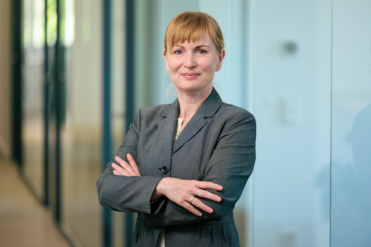 Eine Geschäftsfrau mit einem selbstbewussten Lächeln steht mit verschränkten Armen vor einer Glaswand in einem modernen Bürogebäude. Sie trägt einen grauen Anzug und hat blondes, schulterlanges Haar.