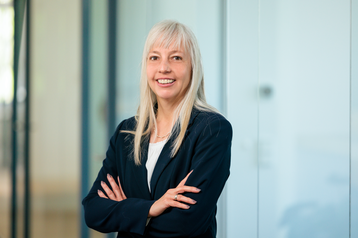 Eine Frau mit langen, blonden Haaren steht mit verschränkten Armen und lächelt in einem modernen Büroumfeld. Sie trägt einen dunklen Blazer und hat einen professionellen, freundlichen Ausdruck.