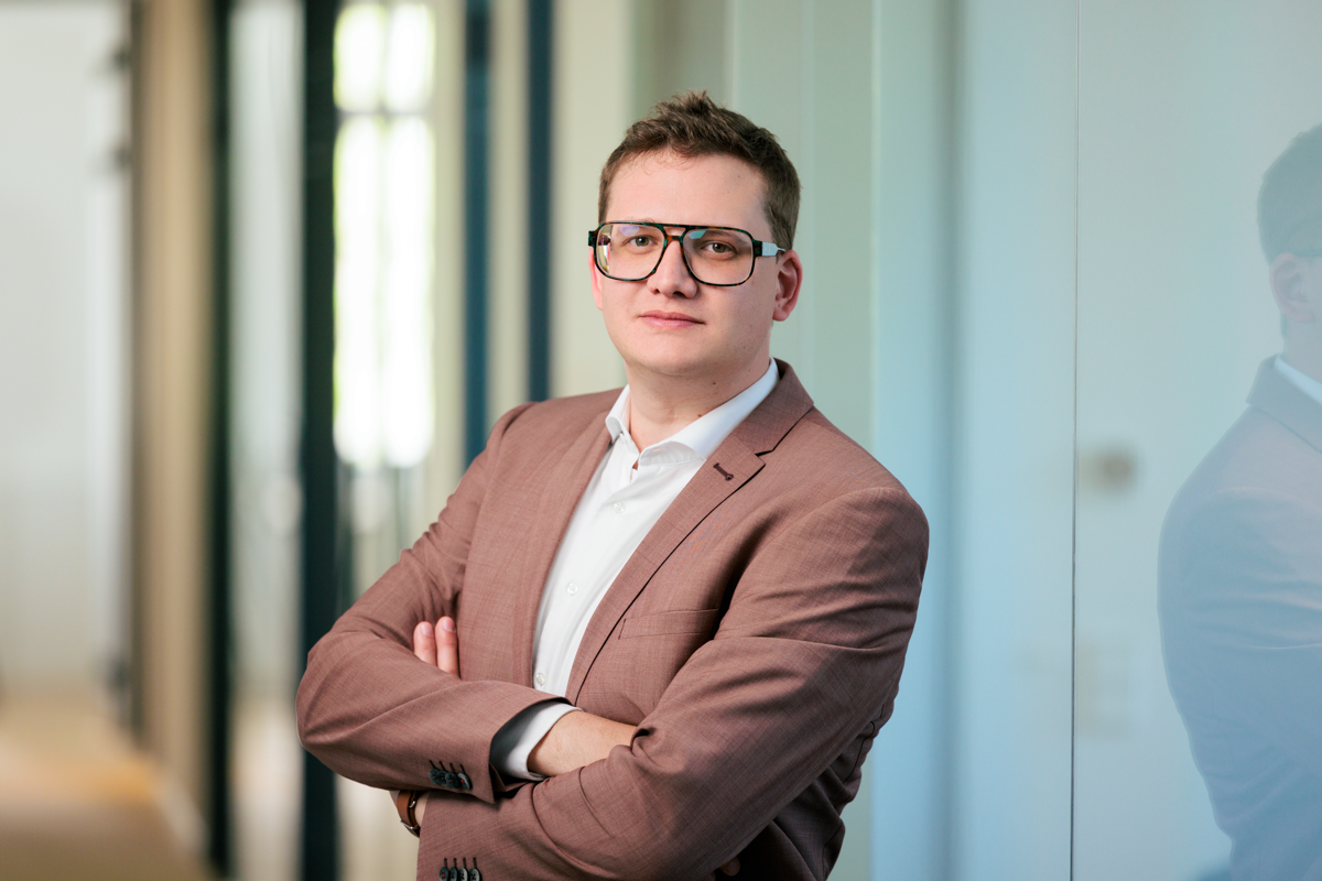 Ein junger Mann mit Brille in einem eleganten braunen Anzug steht mit verschränkten Armen in einem modernen Büro. Er wirkt selbstbewusst und schaut direkt in die Kamera.