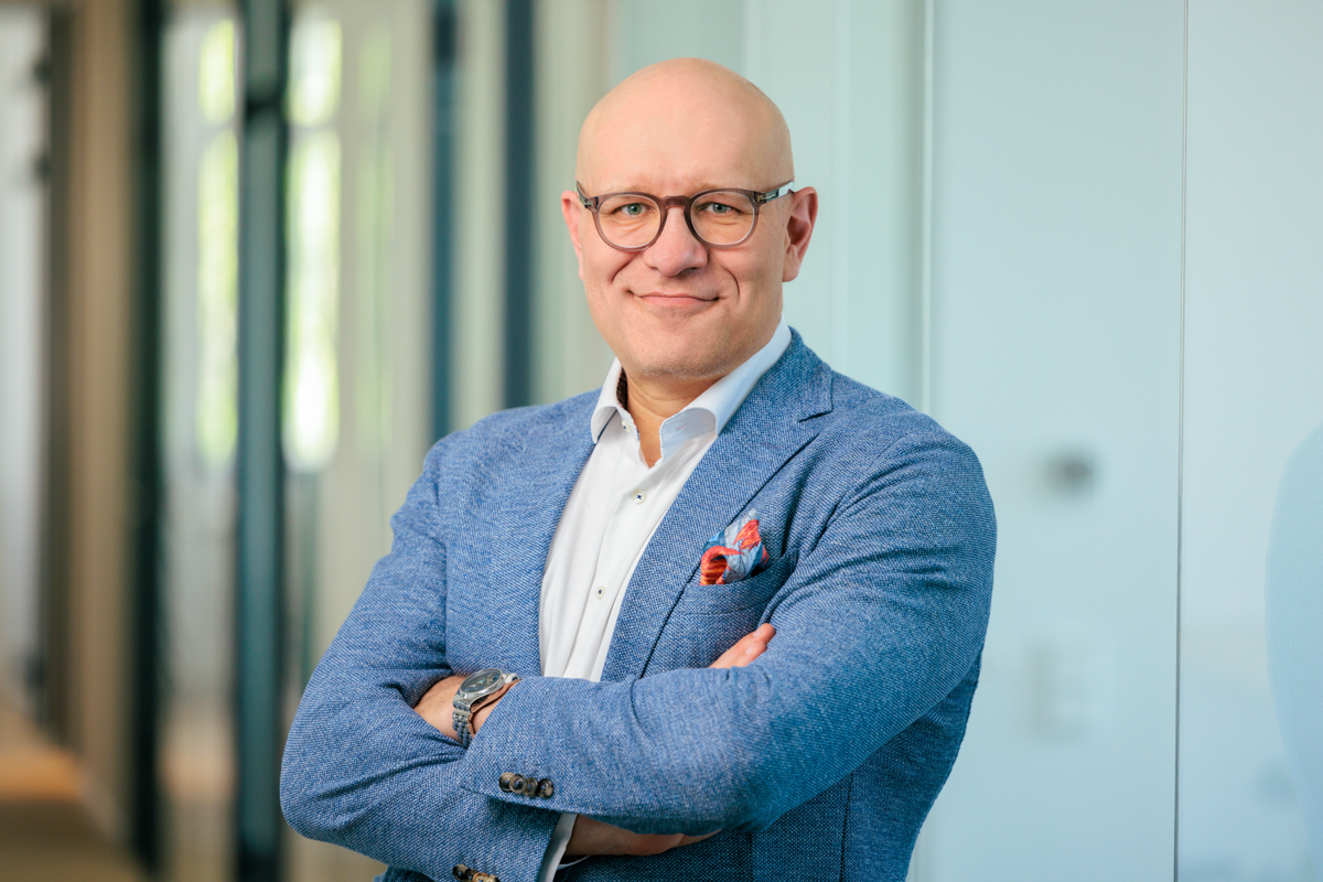 Ein lächelnder Mann mit Glatze und Brille steht mit verschränkten Armen in einem modernen Büro, gekleidet in ein hellblaues Sakko und ein weißes Hemd, mit einem bunten Taschentuch in der Brusttasche.