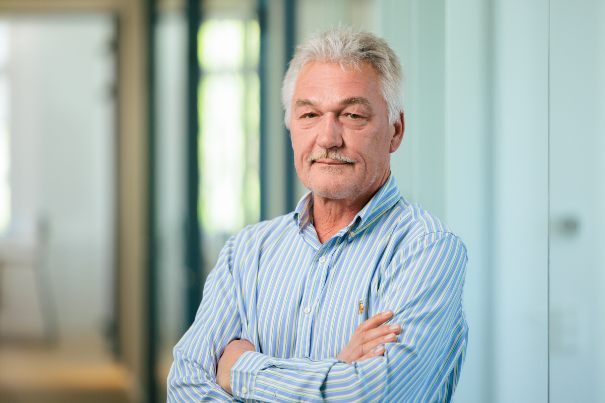 Ein älterer Mann mit grau meliertem Haar und einem Schnurrbart steht mit verschränkten Armen in einem modernen Büroflur. Er trägt ein gestreiftes Hemd und schaut direkt in die Kamera. Die Wände im Hintergrund sind hell und geben einen klaren, professionellen Eindruck.