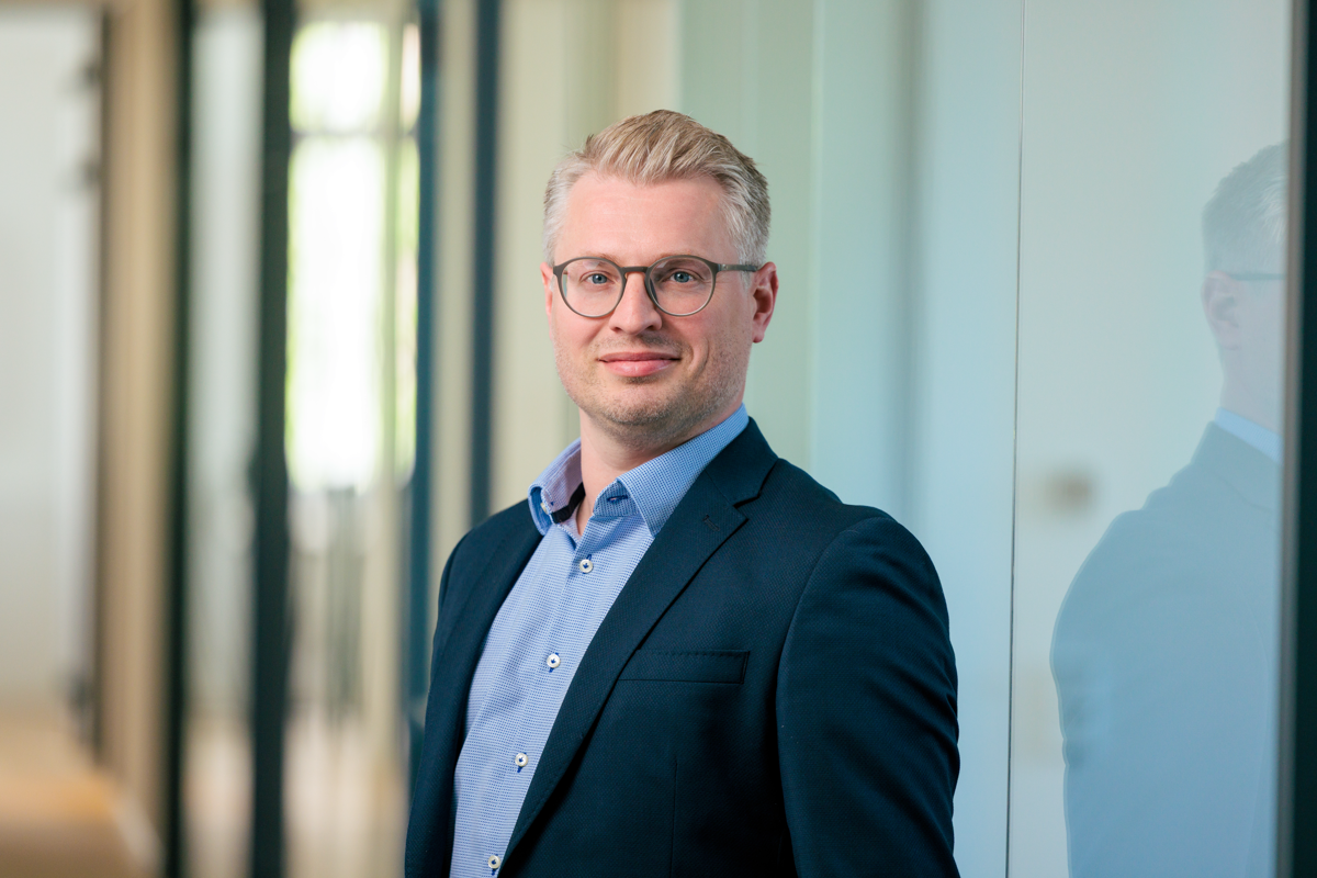 Ein Mann mit Brille und kurzem, blonden Haar steht in einem modernen Büro. Er trägt ein dunkles Sakko und ein hellblaues Hemd und lächelt leicht in die Kamera. Im Hintergrund sind unscharfe Büroelemente zu sehen.