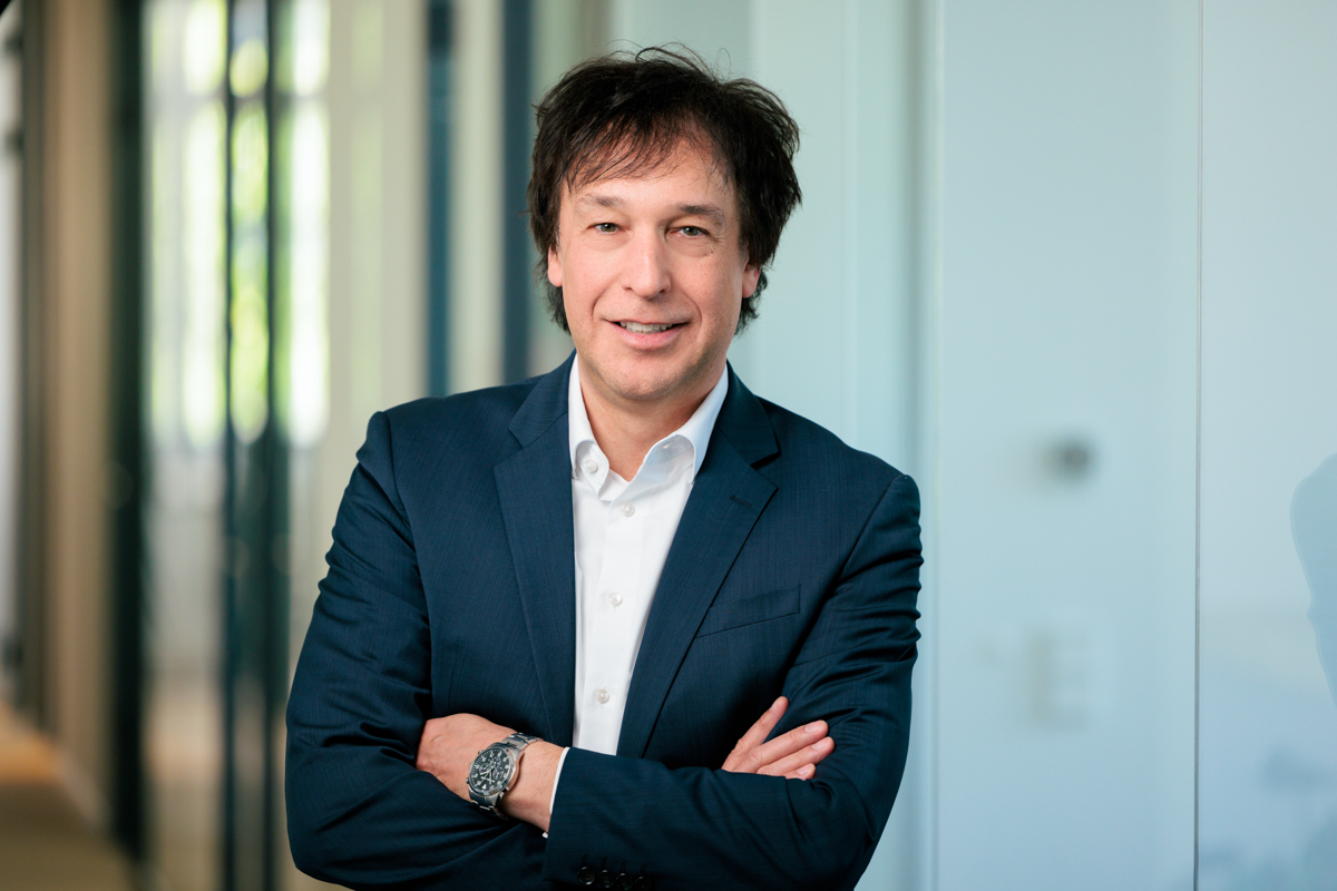 Ein Mann mit dunklem, kräftigem Haar in einem eleganten, dunkelblauen Anzug steht lächelnd mit verschränkten Armen vor einer modernen Büroarchitektur. Das Licht fällt durch große Fenster und schafft eine helle, freundliche Atmosphäre.