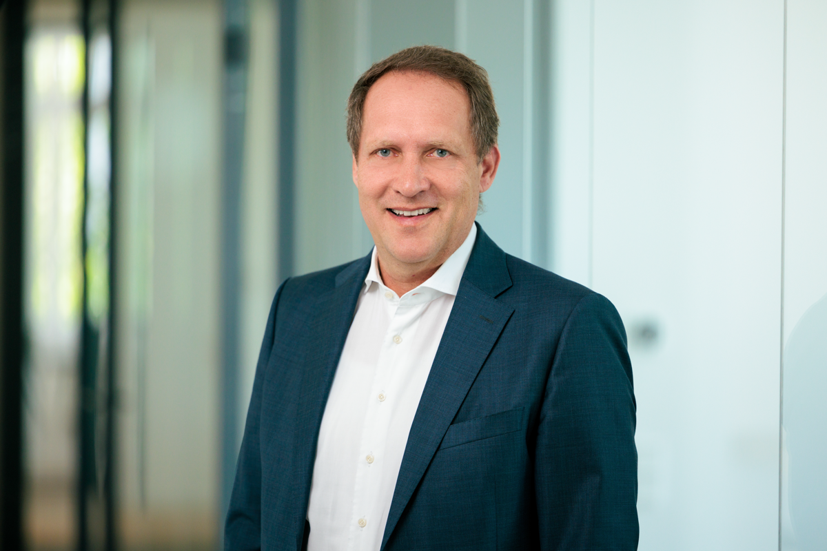 Ein Mann in einem dunkelblauen Anzug und weißem Hemd lächelt freundlich in die Kamera, vor einem modernen, hellen Hintergrund.