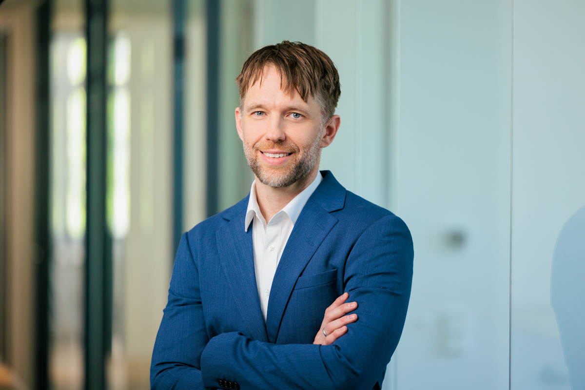 Ein lächelnder Mann mit kurzen Haaren steht in einem modernen Büro, trägt einen blauen Anzug und hat die Arme vor der Brust verschränkt. Im Hintergrund sind verschwommene Fenster und Wände sichtbar.