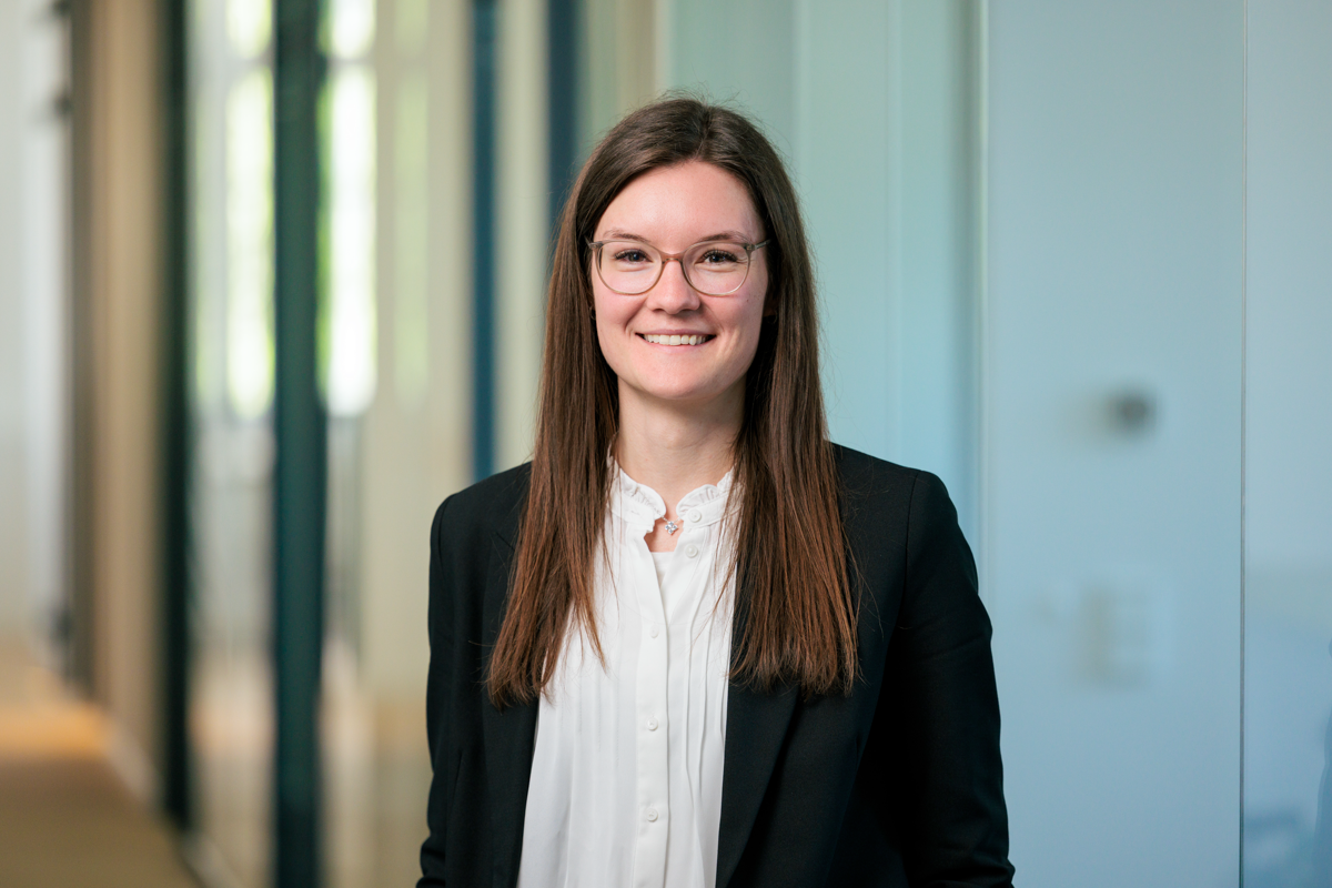 Eine Person mit langen, braunen Haaren und Brille steht vor einer hellen, modernen Büroumgebung und lächelt freundlich in die Kamera. Sie trägt ein schwarzes Blazer und eine weiße Bluse.
