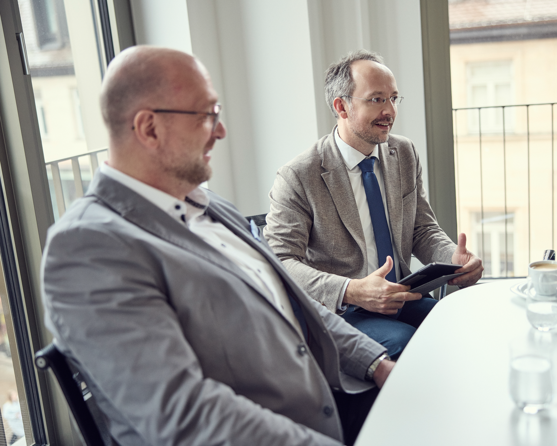 Zwei Männer sitzen an einem Tisch in einem hellen, modernen Büroraum; der Mann rechts spricht und hält ein Tablet in der Hand, während der Mann links aufmerksam zuhört.