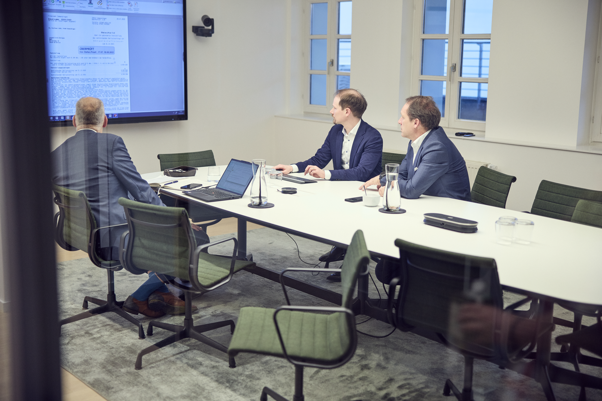 Drei Personen sitzen an einem Konferenztisch in einem hellen Besprechungsraum und schauen auf einen großen Bildschirm, auf dem ein Dokument angezeigt wird. Zwei der Personen sind in Anzügen gekleidet, während eine Person von hinten sichtbar ist und in einem karierten Anzug sitzt. Auf dem Tisch stehen Laptops und Wasserkrüge.