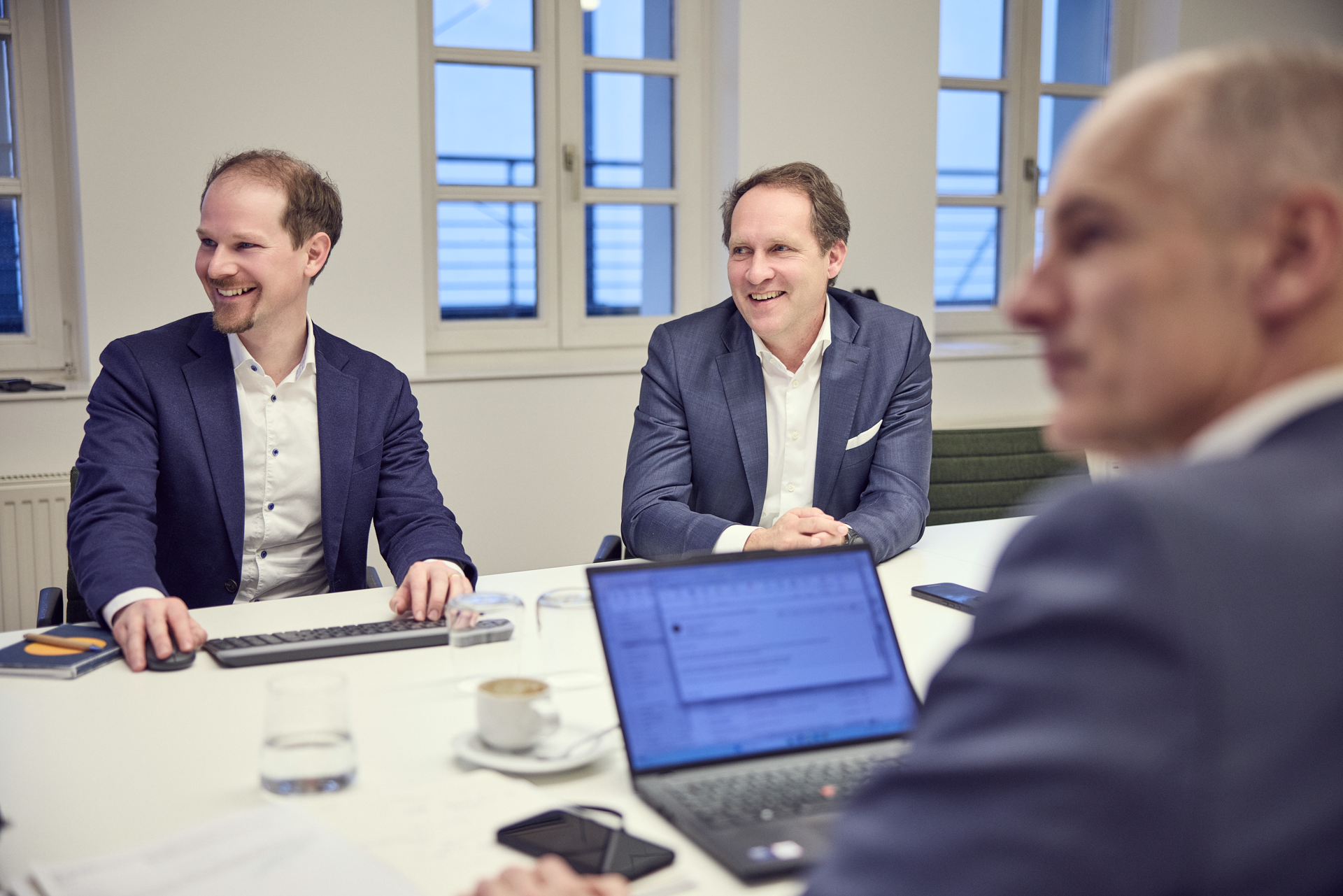 Zwei Männer in formeller Kleidung sitzen an einem Tisch in einem Besprechungsraum und lächeln, während ein dritter Mann in einem Anzug von der Seite auf einen Laptop schaut. Auf dem Tisch stehen Gläser mit Wasser und eine Tasse, während auf dem Laptop ein Dokument sichtbar ist. Durch die Fenster scheint Tageslicht.