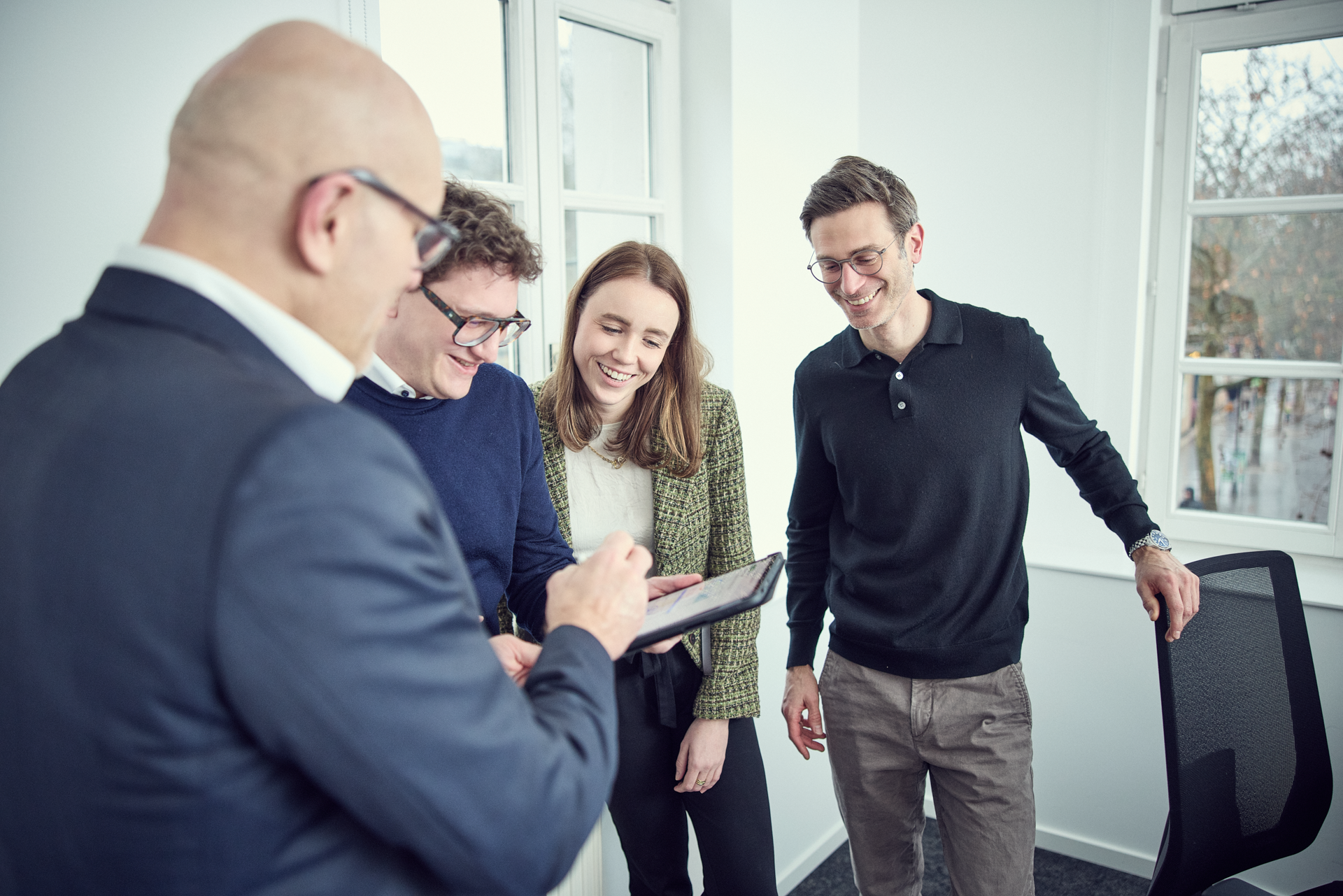 Eine Gruppe von vier Personen in einem hellen Büroraum schaut auf ein Tablet, während sie miteinander sprechen und lächeln.