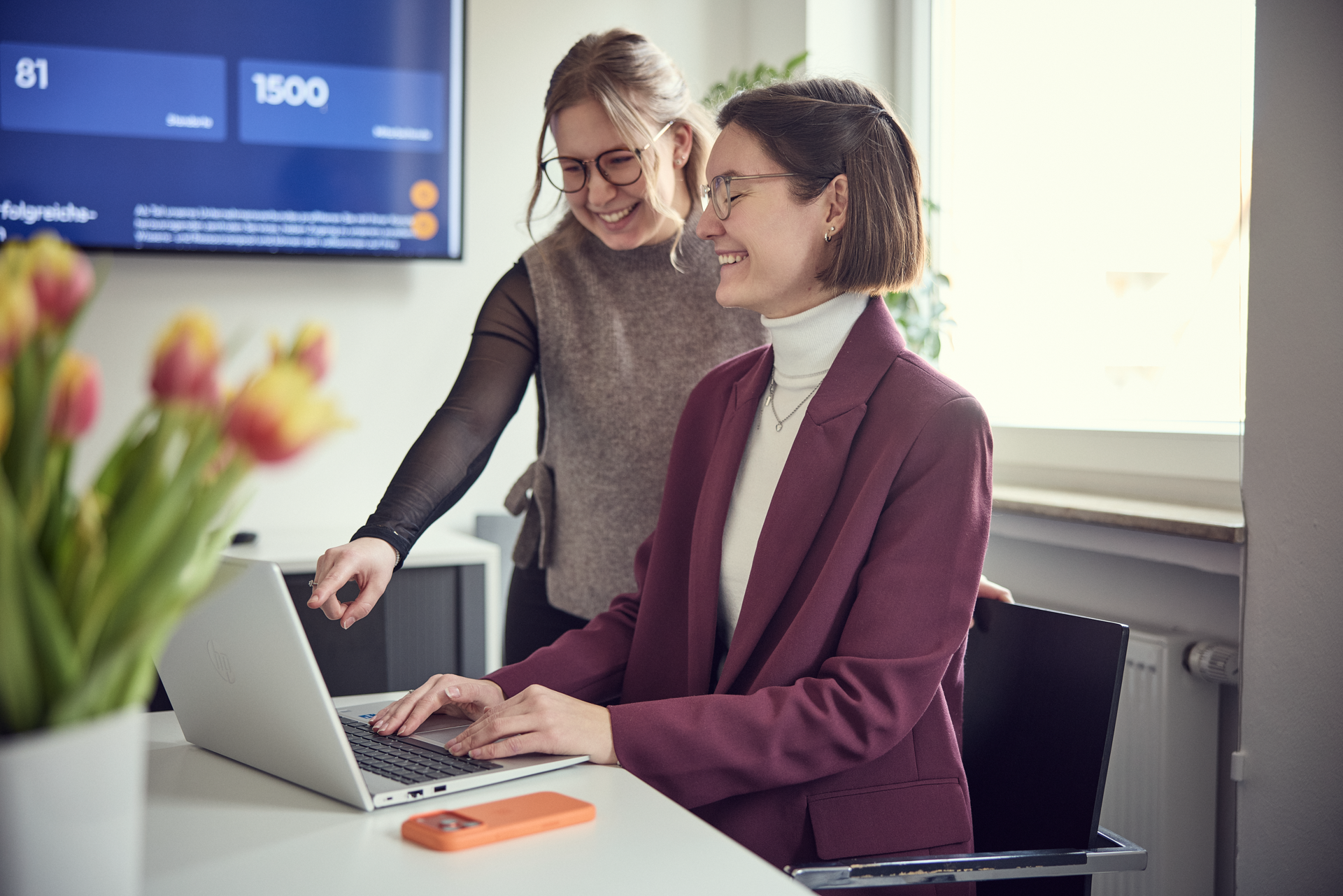 Zwei Frauen sitzen an einem Tisch in einem modernen Büro; eine von ihnen zeigt auf den Bildschirm, während die andere am Laptop arbeitet und lächelt. Auf dem Tisch liegen bunte Tulpen und ein Smartphone.