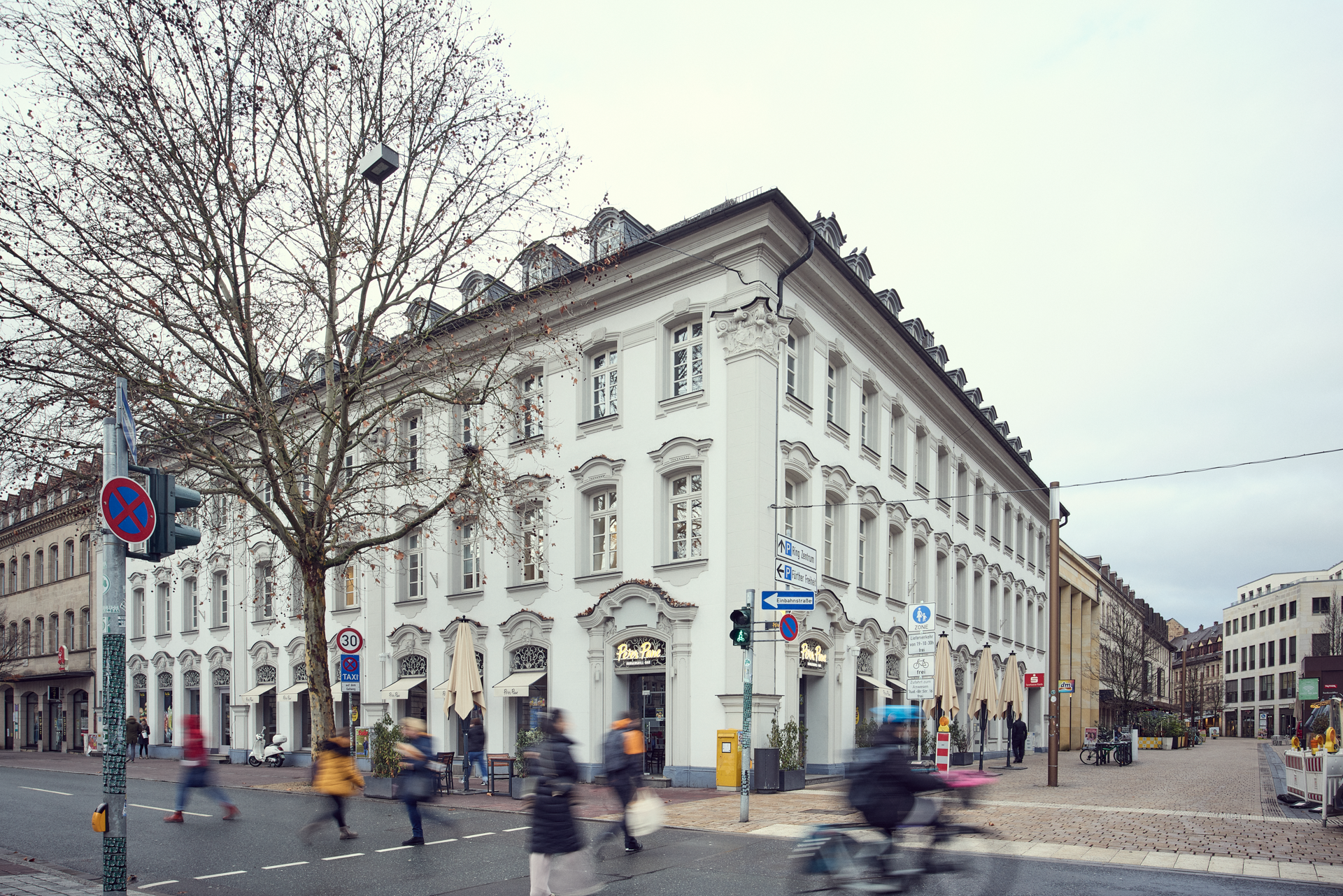 Ein elegantes, historisches Gebäude mit weißer Fassade und vielen Fenstern an einer Straßenecke, umgeben von Fußgängern und modernen Verkehrsschildern. Im Vordergrund sind unscharfe Passanten zu sehen, die die Straße überqueren, während die Bäume im Hintergrund verblasst sind.