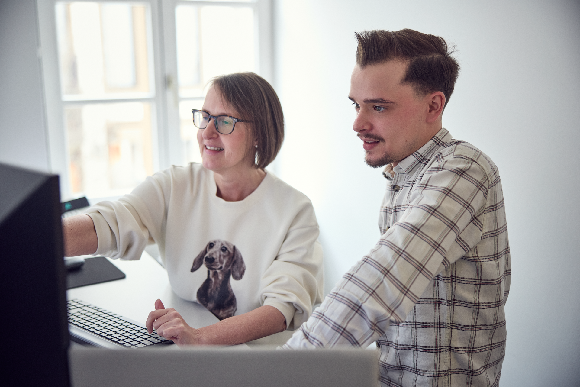 Eine Person mit einer Brille und einem Hundepullover zeigt auf den Bildschirm, während eine andere Person in einem karierten Hemd interessiert zuschaut. Beide stehen an einem Schreibtisch in einem hellen Raum mit Fenster.